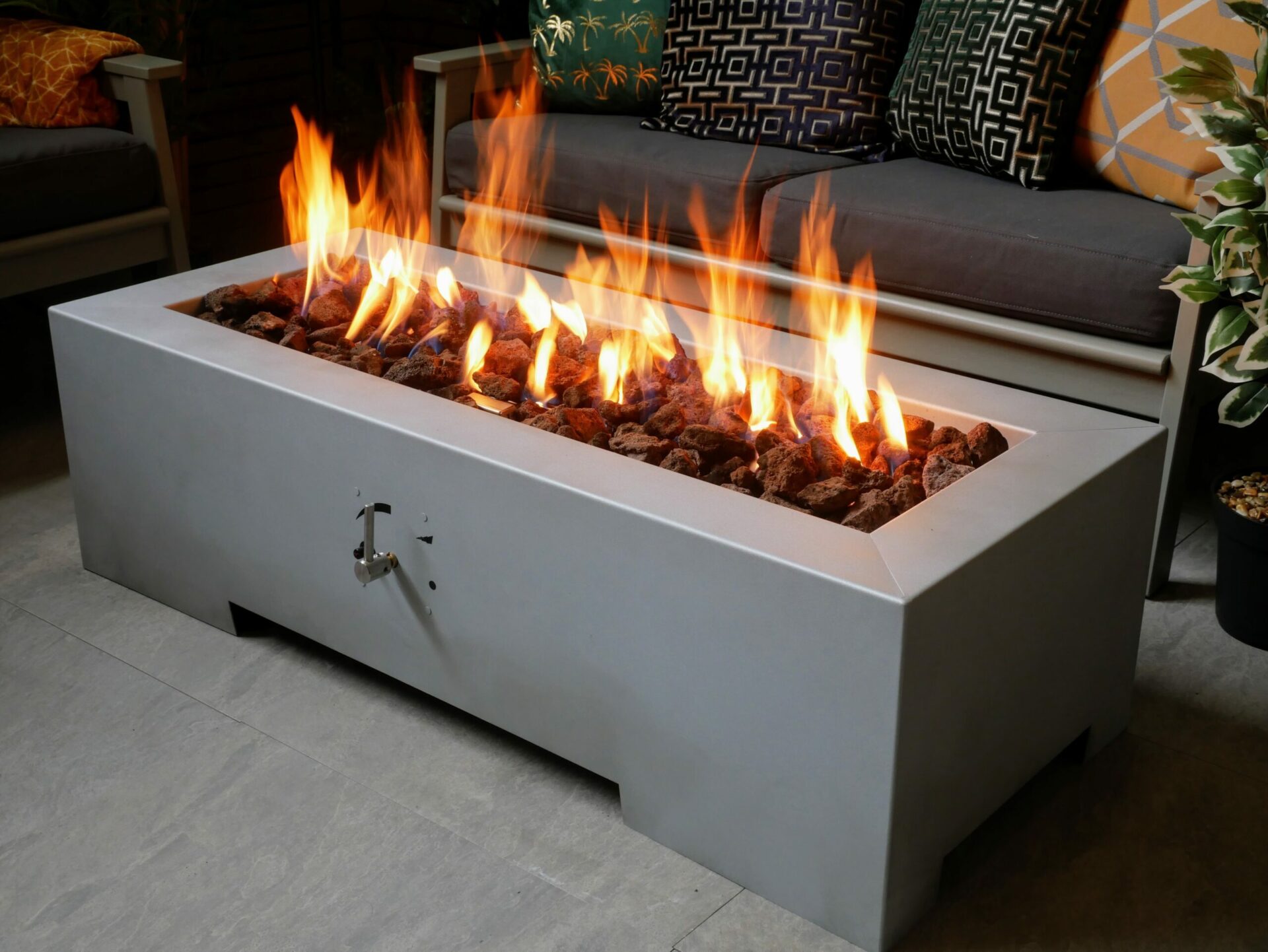 A long rectangular fire pit with flames, set against an outdoor seating area with decorative cushions.